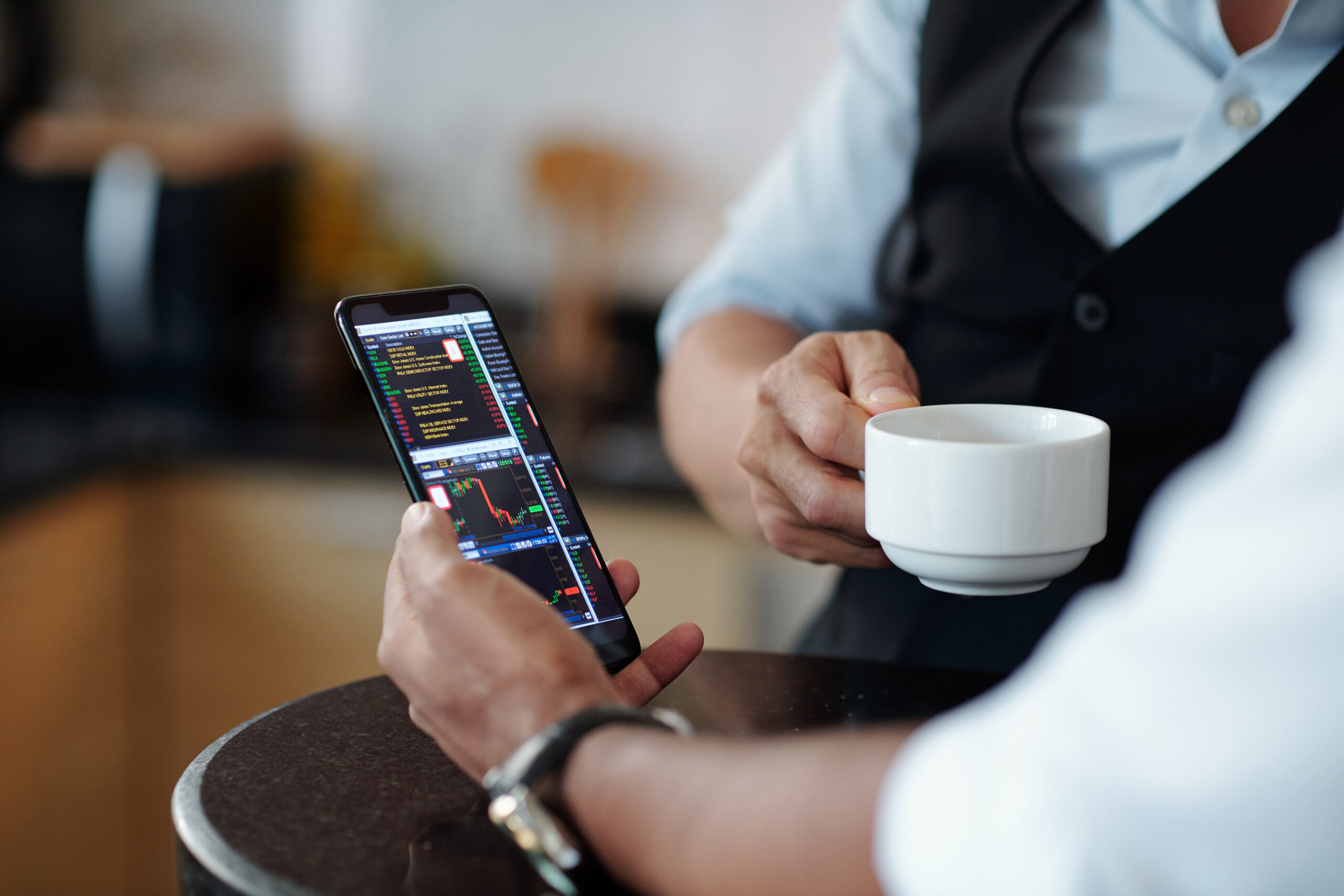 A business professional holding a phone with the screen displaying trading strategies, showcasing his focus on financial decisions, while another professional holds a cup of coffee, symbolizing a blend of productivity and relaxation in the business world.