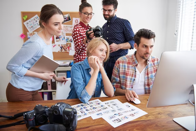 A photo depicting coworkers in an office setting, some holding photo cameras and others working on a computer, showcasing a collaborative and creative work environment.