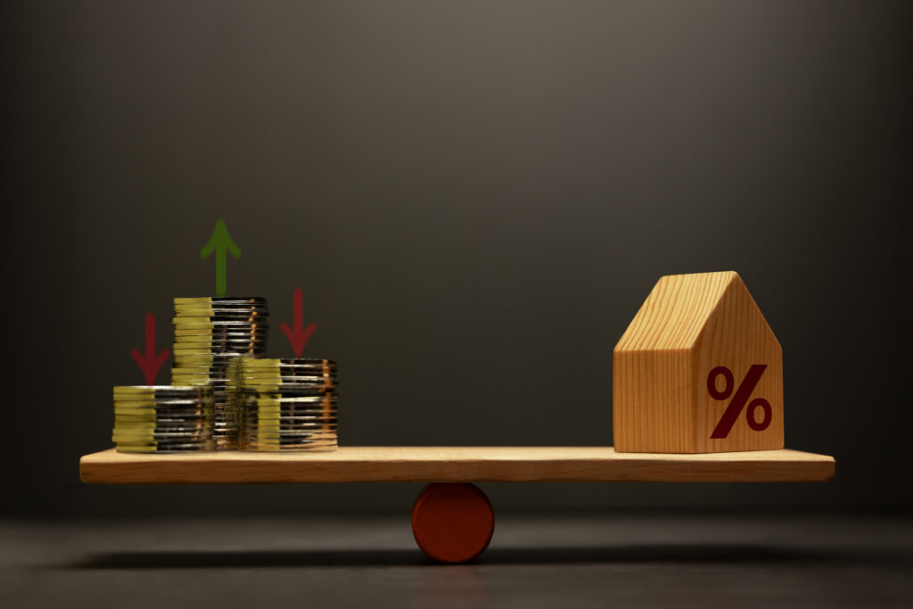 Stacked coins and house balanced on a seesaw scale with percentage symbol representing fixed lot vs percentage risk trading strategy.
