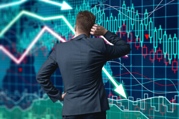 A businessman in a suit from behind, scratching his head while looking at a complex digital screen displaying fluctuating stock market candlestick charts and downward-pointing trend arrows.