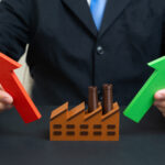 A person in a suit holding a red upward arrow and a green upward arrow on either side of a miniature brown factory model, symbolizing industrial growth and market expansion.