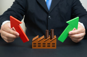 A person in a suit holding a red upward arrow and a green upward arrow on either side of a miniature brown factory model, symbolizing industrial growth and market expansion.