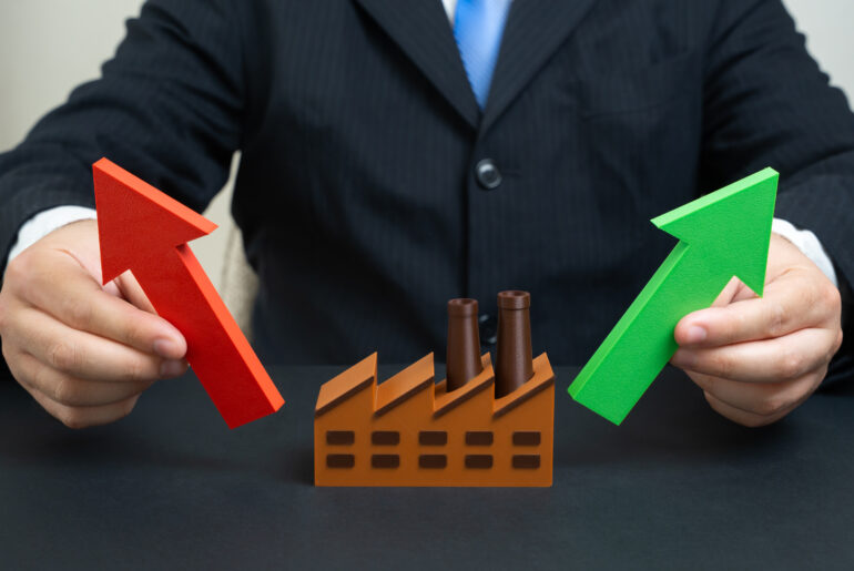 A person in a suit holding a red upward arrow and a green upward arrow on either side of a miniature brown factory model, symbolizing industrial growth and market expansion.