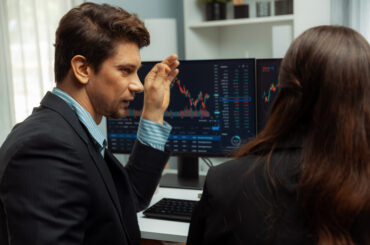 A male forex trader pointing at a computer screen showing financial candlestick charts while explaining market concepts to a colleague.