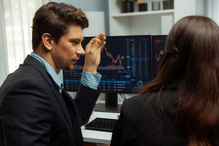 A male forex trader pointing at a computer screen showing financial candlestick charts while explaining market concepts to a colleague.