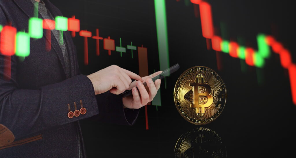 A double-exposure image of a professional in a dark blazer using a smartphone. In the background, a glowing red and green candlestick chart trends downward, while a large, physical-style gold Bitcoin coin floats in the foreground with a clear reflection.