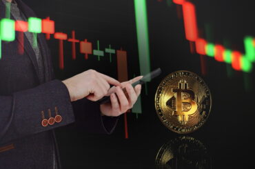 A double-exposure image of a professional in a dark blazer using a smartphone. In the background, a glowing red and green candlestick chart trends downward, while a large, physical-style gold Bitcoin coin floats in the foreground with a clear reflection.