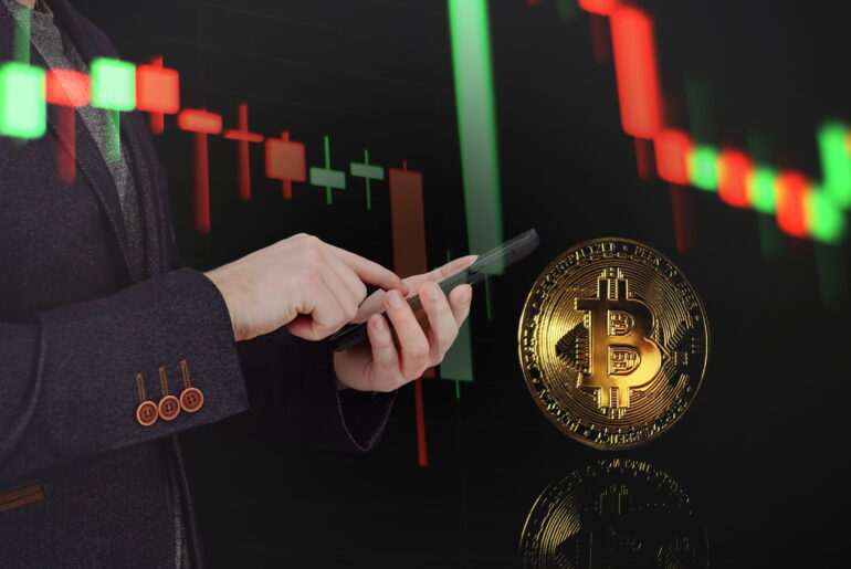 A double-exposure image of a professional in a dark blazer using a smartphone. In the background, a glowing red and green candlestick chart trends downward, while a large, physical-style gold Bitcoin coin floats in the foreground with a clear reflection.