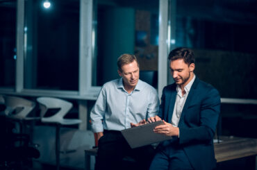 Two corporate professionals in a modern, dimly lit office looking at a tablet screen together, illustrating a discussion on advanced financial market concepts and institutional trading strategies.