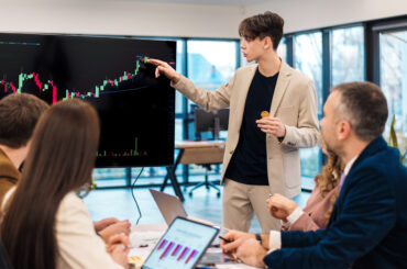 A young financial analyst pointing to a large monitor displaying a bullish candlestick chart during a meeting with a team of traders.