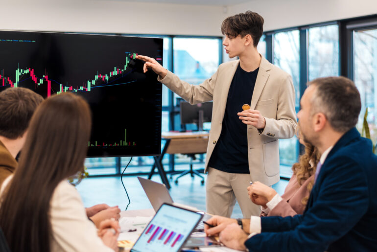 A young financial analyst pointing to a large monitor displaying a bullish candlestick chart during a meeting with a team of traders.