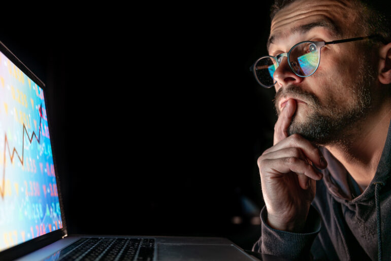 A close-up of a trader with glasses thoughtfully looking at a laptop screen showing a rising financial stock chart in a dark room.