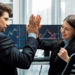 Two professional traders giving a high five in front of stock market charts, celebrating a successful trade executed using objective risk management rules.