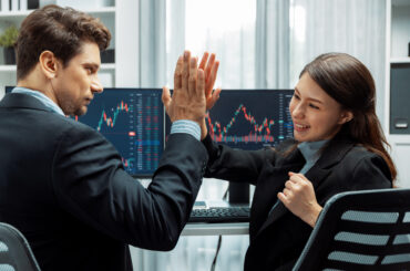Two professional traders giving a high five in front of stock market charts, celebrating a successful trade executed using objective risk management rules.