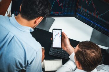 Trader analyzing dynamic candlestick price charts on MetaTrader 5 using a smartphone and dual desktop monitors.