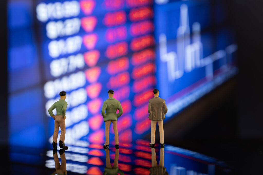 Miniature figures of financial traders standing in front of a blurred stock exchange display board showing market volatility during a recession.