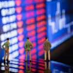 Miniature figures of financial traders standing in front of a blurred stock exchange display board showing market volatility during a recession.