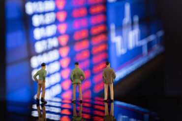 Miniature figures of financial traders standing in front of a blurred stock exchange display board showing market volatility during a recession.