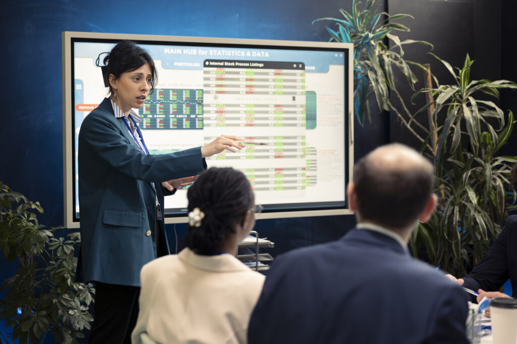 A female market analyst pointing to an interactive digital screen showing economic data and financial market statistics during a presentation.