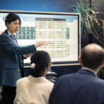 A female market analyst pointing to an interactive digital screen showing economic data and financial market statistics during a presentation.