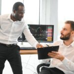 Two diverse colleagues discussing market charts and sharing stock trading strategies in front of multiple computer monitors showing financial price charts.