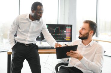 Two diverse colleagues discussing market charts and sharing stock trading strategies in front of multiple computer monitors showing financial price charts.