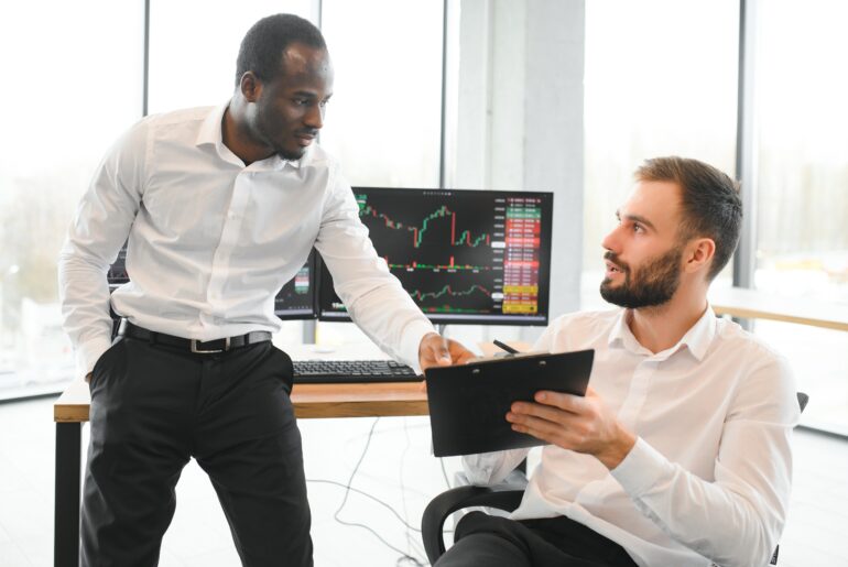 Two diverse colleagues discussing market charts and sharing stock trading strategies in front of multiple computer monitors showing financial price charts.