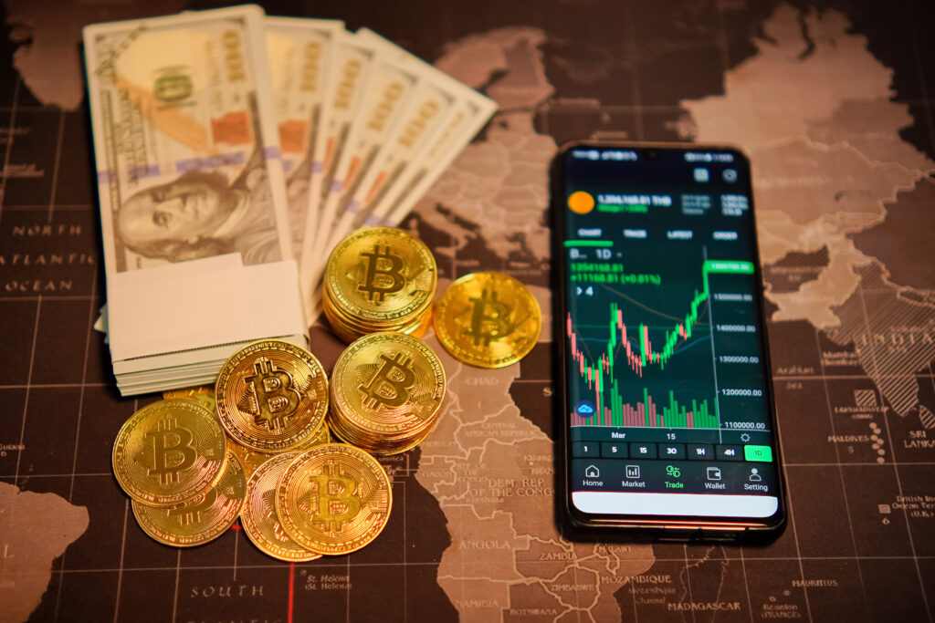 Physical fiat currency cash notes alongside crypto coins and a smartphone displaying a digital asset trading chart on a world map background.