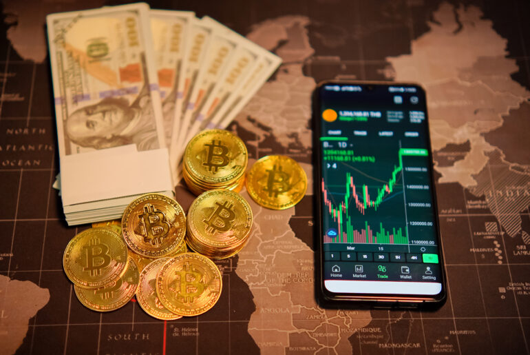 Physical fiat currency cash notes alongside crypto coins and a smartphone displaying a digital asset trading chart on a world map background.
