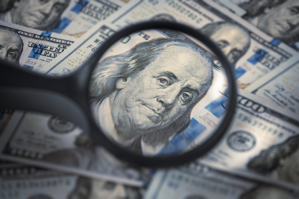 Close-up of a magnifying glass focusing on Benjamin Franklin's portrait on a 100 US dollar bill over a pile of USD currency.