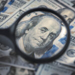 Close-up of a magnifying glass focusing on Benjamin Franklin's portrait on a 100 US dollar bill over a pile of USD currency.